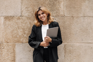 Smiling beautiful woman looking at camera in hands laptop. Wearing black business costume staying...