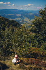 Fototapeta na wymiar A little girl in a pink hat sits on the grass in the mountains, hot summer. Around the trees and blue sky with clouds.