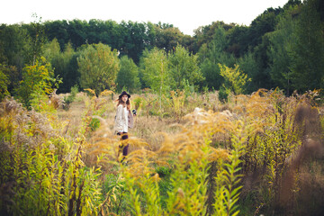 Obraz premium A stylish young girl photographer with blond hair, in a hat, a light jacket and burgundy pants, with a camera, walks through a field with tall grass, on a bright sunny evening in summer.