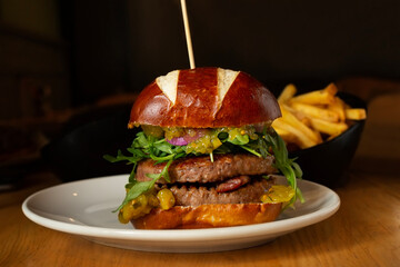 Fast food concept. Pretzel bread burger with bacon, rocket salad, relish, cucumber, sauce, onion. French fries, dark restaurant background.