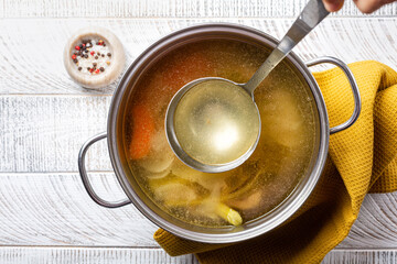 Cooking beef broth stock in a saucepan with a ladle , made using meat, bones, vegetables, carrot, onin, celery, and seasoning. - obrazy, fototapety, plakaty