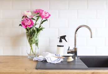 Peonies flowers in a vase on a wooden table. Blurred kitchen background. Contemporary elegant scandi interior. Set of cleaning products on a light background. White plastic pump bottle for shampoo and