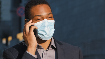 African American businessman ethnic man male guy in medical face mask talking mobile phone answer call talk with smartphone calling speaking with cellphone in city outdoors coronavirus covid pandemic