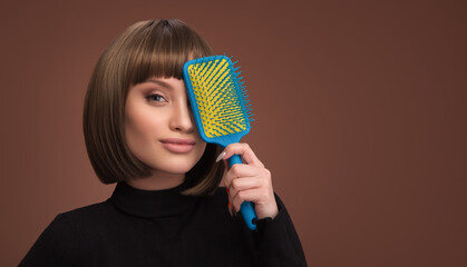 A young smiling girl with a bob haircut holds a comb in her hand. Brown background.