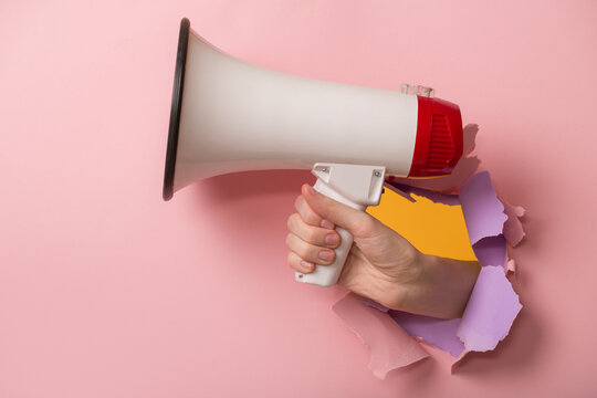 A hand holds a megaphone through a hole in pink paper.
