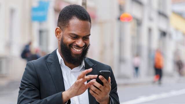 Smiling ethnic African American happy bearded man businessman executive employer with smartphone in city chatting texting has business chat reading good news online using mobile app browsing phone