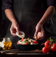 Cooking process of Greek Salad for a restaurant menu. Food composition of a Mediterranean cuisine in rustic style. Chef put the onion and olives in a black bowl and mixed of all vegetables in a salad.