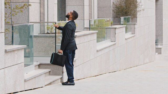 African American ethnic man candidate to job offer work interview standing near office building looking at wristwatch. Businessman entrepreneur hurry to business meeting check time standing in city