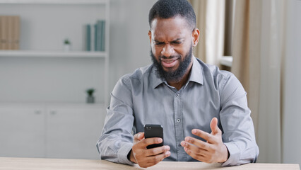 Angry African American ethnic bearded man boss businessman with mobile phone smartphone mistake failure reading bad news refuse notice mad with business trouble problem sitting at table in office