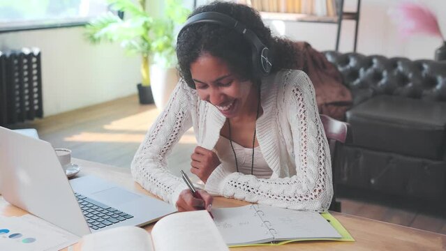 Young happy African American woman in headphones listens to internet podcast and laughs making notes in workbook gets quality education and opportunity to change careers sits at table in home interior