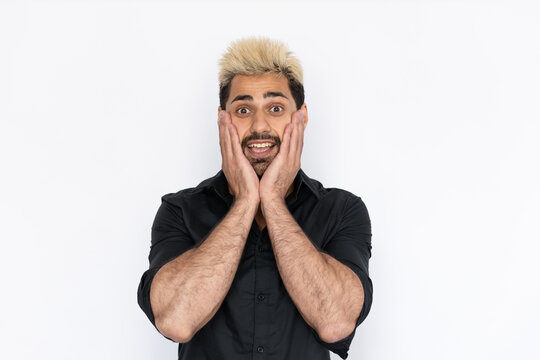 Surprised young man holding his face with hands from shock. Male Caucasian model with brown eyes, ombre painted hair and beard in black shirt touching his face from surprise. Amazement concept