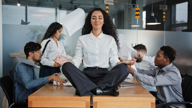 Calm businesswoman female leader woman sitting on table in lotus position meditating in office feels calmness ignore annoyed angry colleagues throw documents business conflict quarrel on background