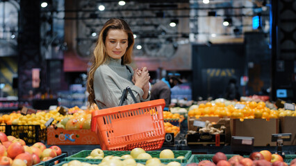 Fototapeta Caucasian female consumer woman shopper buyer with basket walks to grocery store supermarket looking for goods looking at stalls with fresh tasty tropic fruits buying food making choice discount sale obraz