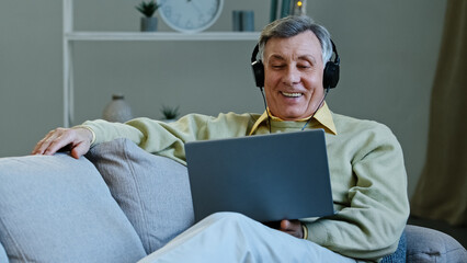 Excited happy aged man watching movie using laptop and headphones sitting lying on comfy couch smiling elderly grandfather relaxing watching comedy show on modern device laughs happily enjoy free time