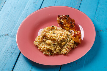 Couscous with vegetables in sauce and spices with grilled chicken drumstick on a pink plate on a blue table.