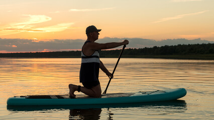 A man on his knees on a SUP board with an oar at sunset swims in the calm water of the lake against...
