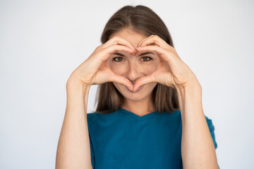 Happy Caucasian woman showing heart gesture. Portrait of pleased young female model with brown hair in blue T-shirt looking at camera, smiling behind heart shape. Love, romance concept