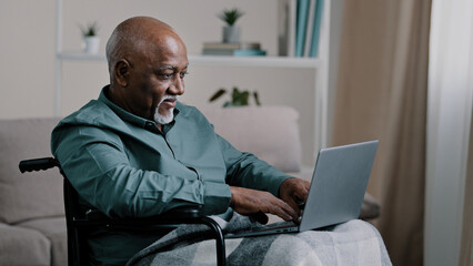 Old 60s african business man in wheelchair indoor at home typing on laptop communicates remotely working online distant texting chatting with family grandfather with disability use modern computer app