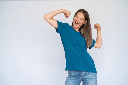 Excited Caucasian woman showing biceps. Joyful young female model with brown hair in blue T-shirt with closed eyes laughing, dancing with raised arms. Optimism, success concept