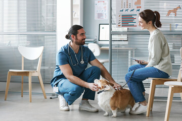 Young veterinarian doctor in blue uniform talking to owner of pet