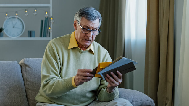 Old 60s elderly Caucasian man checking postal letters at home. Mature male in living room senior grandfather sitting on sofa looking at correspondence bank notification pensioner check bills indoors