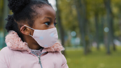 Close up African American small girl ethnic baby pupil schoolgirl ill sick daughter child kid in medical face mask looking away to side outdoors in city back to school in coronavirus pandemic covid-19