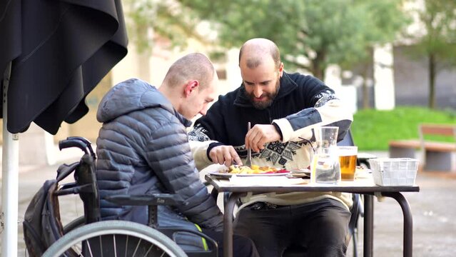 A disabled person eating on the terrace of a restaurant with a friend helping him eat