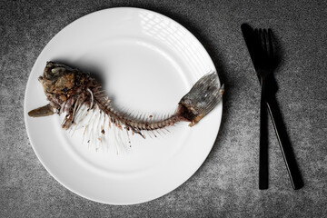 Fried fish skeleton, spine and bones on a white plate with a black knife and fork on a gray background.