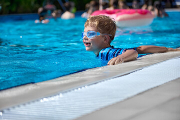 Boy in a bathing suit and goggles swims merrily in a warm outdoor pool with clear clear water. A...