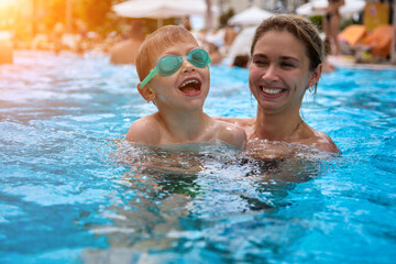 Cute young mother teaches her beloved son to swim in the pool in bathing glasses at the resort in the summer. Woman smiles, swims with her son in an outdoor pool on a summer vacation