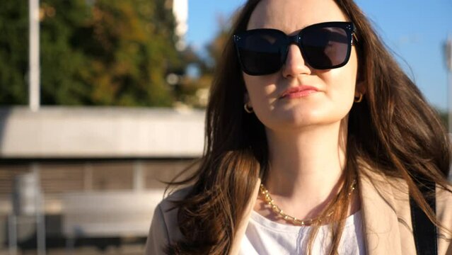 Portrait of beautiful lady in sunglasses walking at sunny day. Attractive brunette girl looks into camera going along city street. Businesswoman commuting to work. Concept of urban lifestyle. Close up