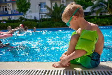 Sad boy in bathing shorts sits near the outdoor pool in summer, they are not allowed to swim. Small child is forbidden to swim in a deep pool for adults