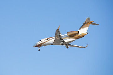 Avion jet privé volant en phase d'atterrissage, trains de roues sortis. Petit avion à réacteurs blanc et doré sur fond de ciel bleu.