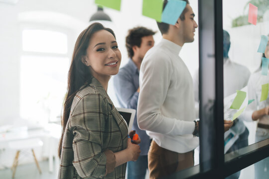 Smiling female employee standing in office with team working on transparent wall