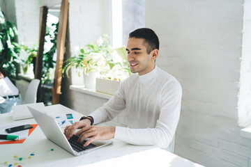 Smiling young man typing on laptop while sitting at table in office