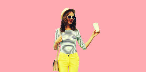 Portrait of happy smiling stylish young woman with coffee cup wearing handbag, striped t-shirt and summer straw round hat on pink background