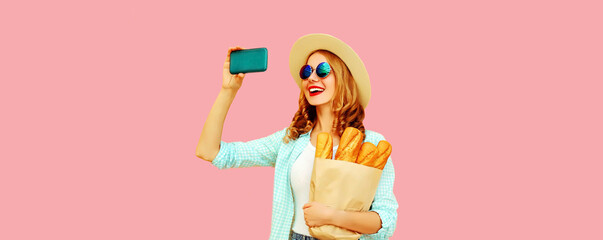 Portrait of happy smiling woman taking selfie with smartphone holding grocery shopping paper bag with long white bread baguette on pink background