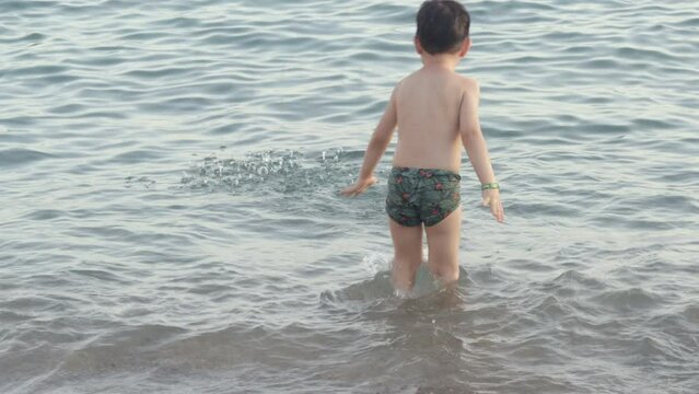 kid baby having fun on beach seashore against sea waves digging in sand jumping splashing water with hands.child boy with blue inflatable ring stay bending on sands,toddler infant no clothes 4k real