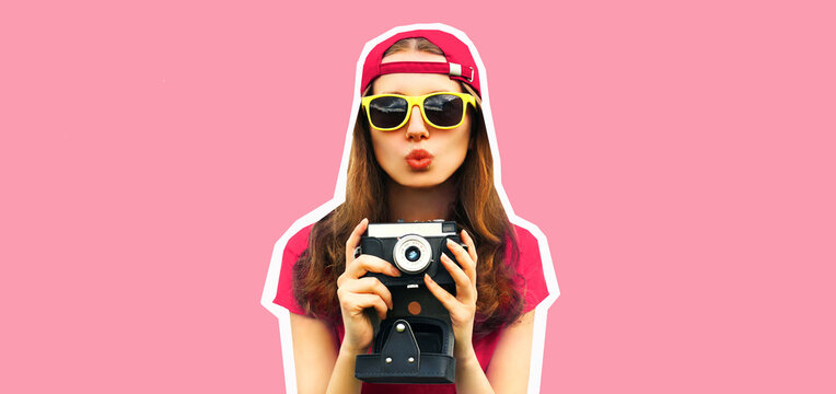Portrait of happy young woman photographer taking picture on film camera and blowing her lips wearing baseball cap on pink background, magazine style