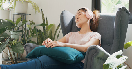 Close-up Young Asian woman with casual wear headphone sit on wing chair close eyes use phone listen calm music relax in living room at home. Cozy house interior indoor plants, Urban jungle concept.