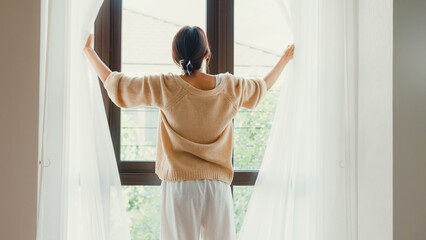 Fototapeta na wymiar Happy Asian girl with white cream pajamas open white curtain on window looking outside feel fresh with peaceful morning and fresh air on weekend in bedroom at home. Female morning lifestyle concept.