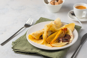 Thin pancakes stuffed with caramelized bananas and chocolate spread on a white plate against a light concrete background. International pancake day.