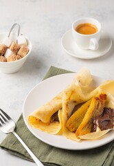 Thin pancakes stuffed with caramelized bananas and chocolate spread on a white plate against a light concrete background. International pancake day.