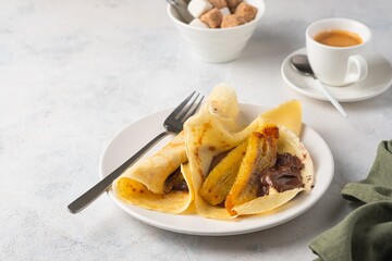 Thin pancakes stuffed with caramelized bananas and chocolate spread on a white plate against a light concrete background. International pancake day.