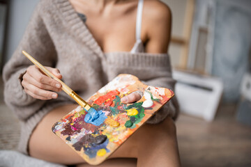 Cropped view of burred artist holding paintbrush and palette in workshop