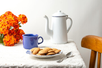 Top view of still life on table with white tablecloth and chair, with cookies, old coffee pot, blue cup and orange flowers, white background, horizontal with copy space