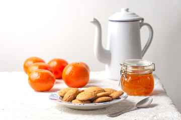 Still life view of biscuits, old coffee pot, jam, spoon and tangerines on white tablecloth, white background, horizontal with copy space