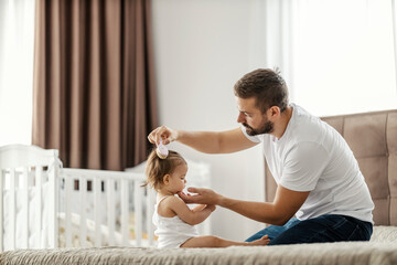 A caring father is brushing his daughter hair and making a hairstyle in a bedroom.