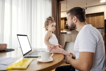 A casual businessman is working from home remotely while taking care of his daughter.