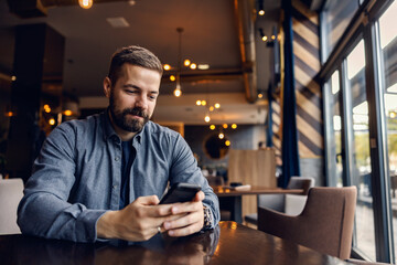 A satisfied man is sitting at the table in coffee shop and scrolling on the phone.